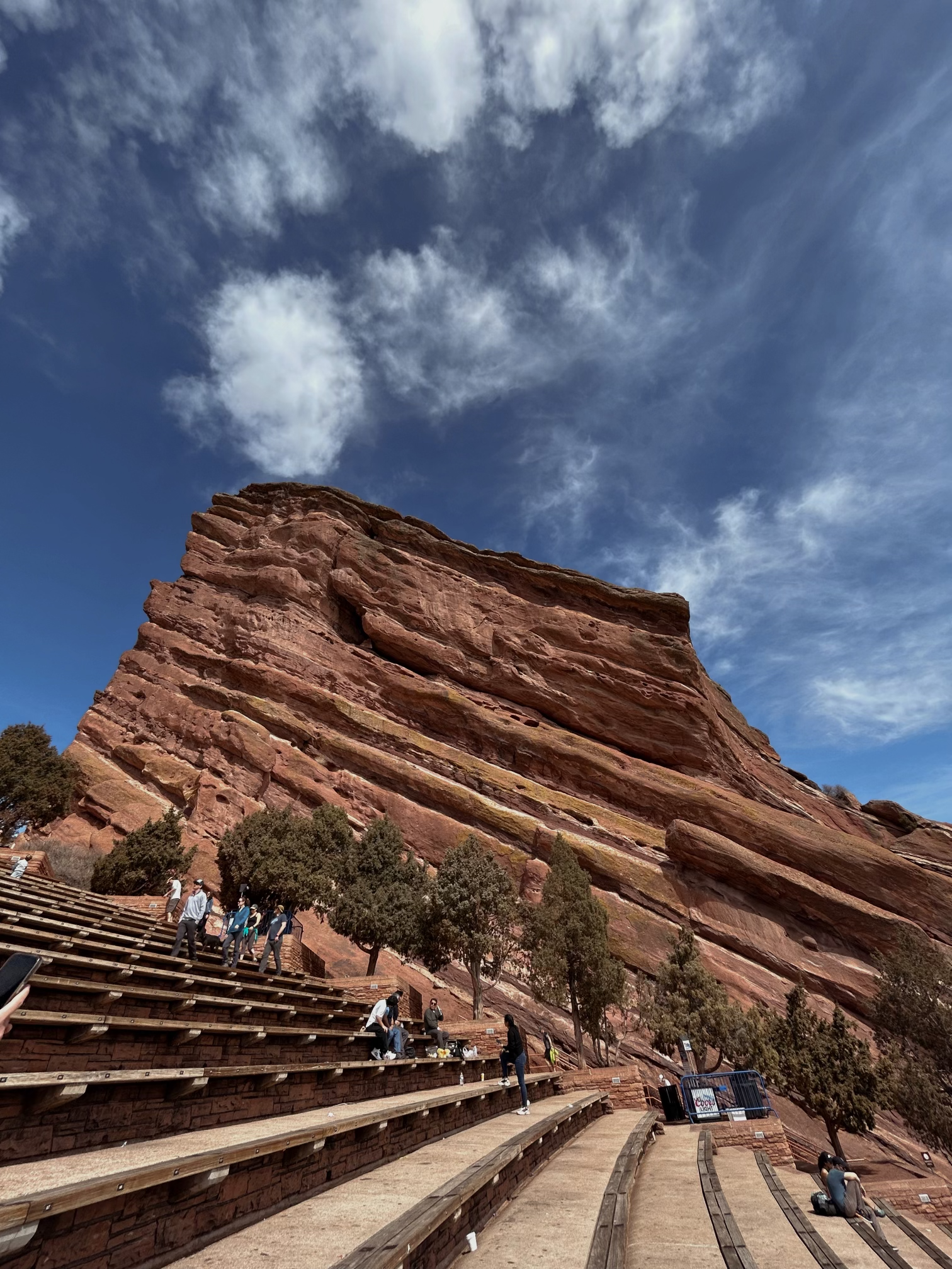 Large redish rock in center of photo. Blue sky with wispy white 
                clouds above. Dry trees in front of rock. Rows of bench seating in foreground
                of picture.