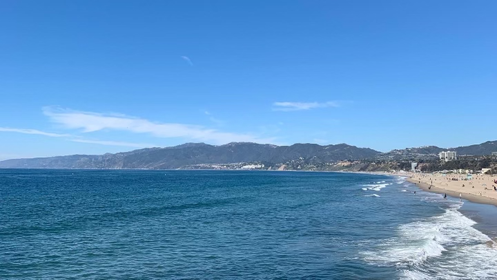 Bright blue ocean in bottom half of picture with beach peaking in 
                to the far right. Mountains in the central horizontal line. Bright blue sky with 
                few clouds.