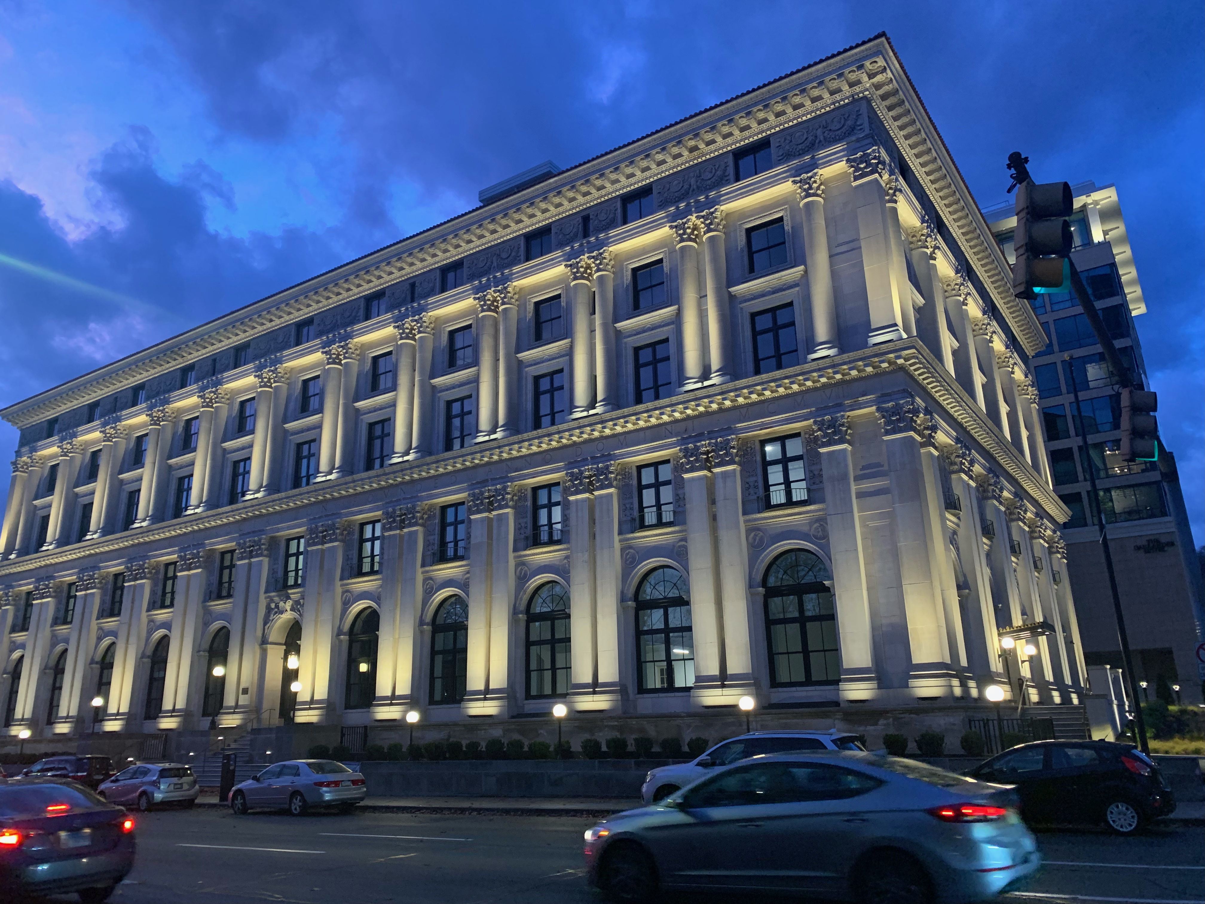 Concrete columned building illuminated from below. Sky is dark; time of day is dusk. Cars
    zoom past on street at bottom of image.