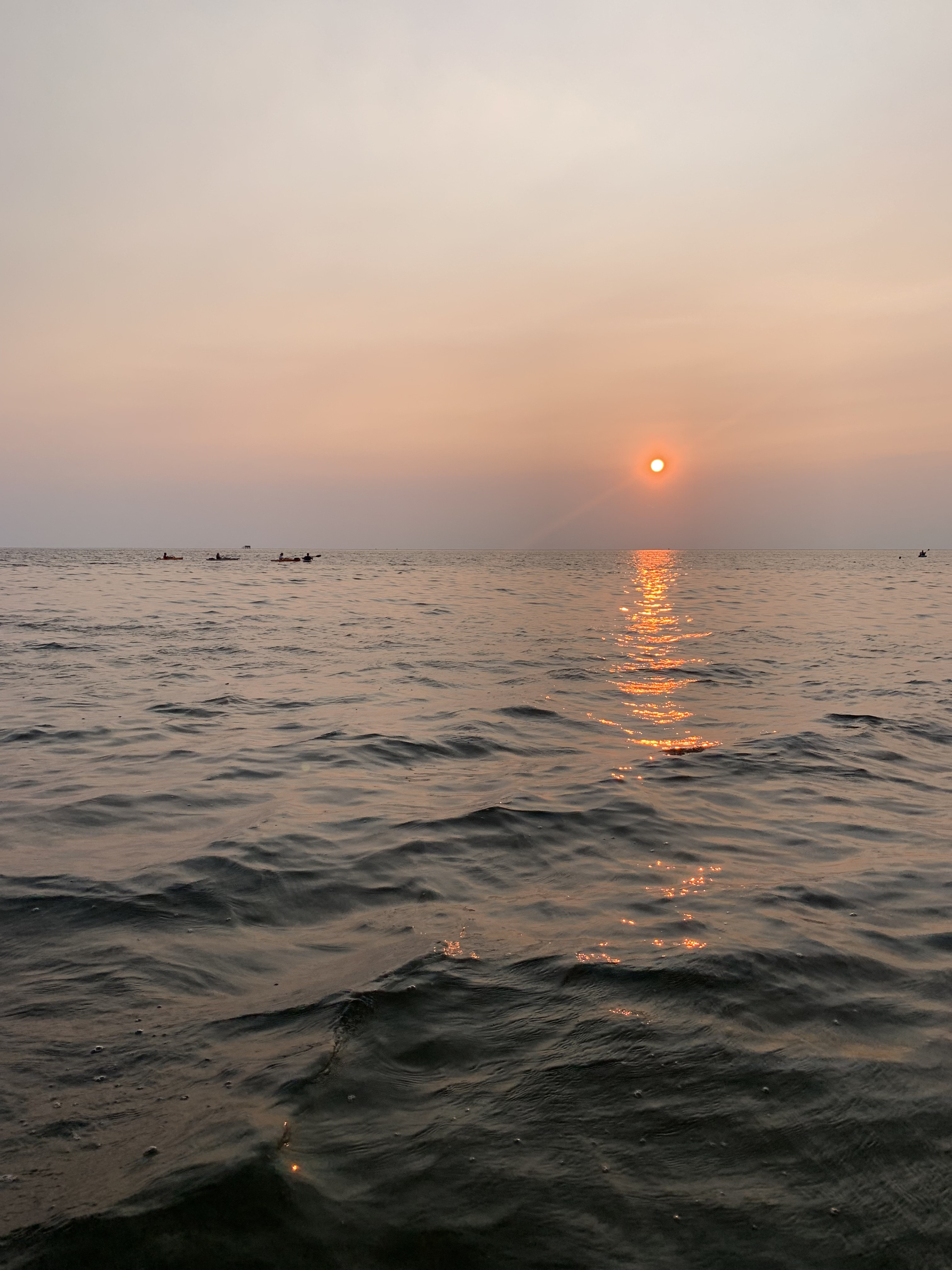 Calm ocean on bottom half of image. Orange sun setting near water line.
                    Sky fading from light purple to peach to gray above the water.