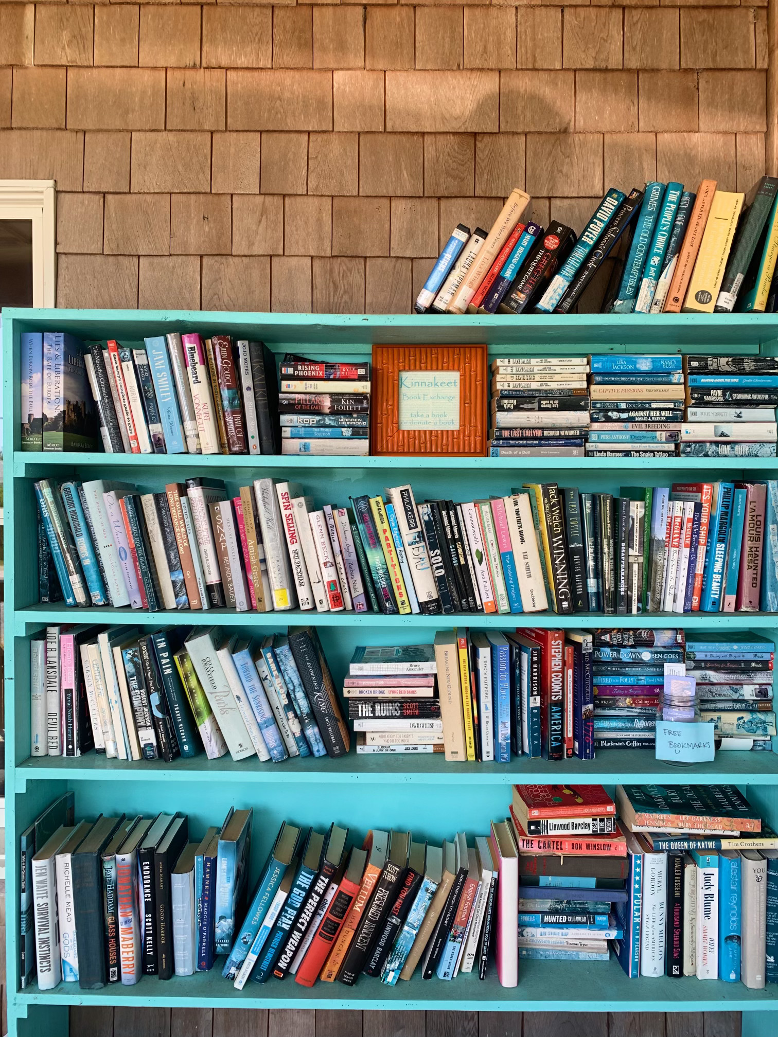 Teal bookshelf filled with stacks of colorful books.
                    Lightly-colored wooden shingles side the building above the bookshelf.