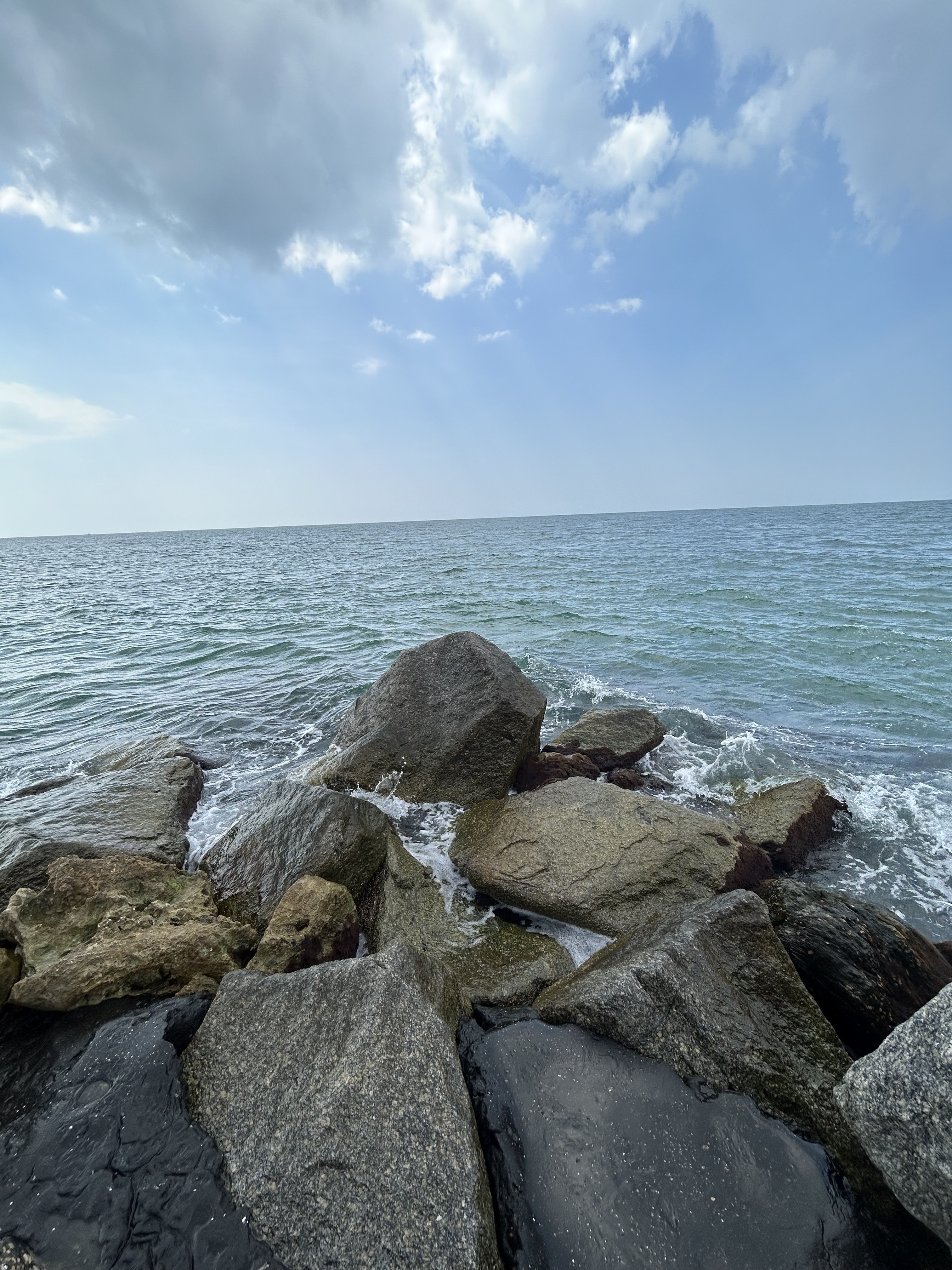 Large rocks protruding out into ocean in triangle-formation.
                Dark teal ocean with small waves. Light blue sky with white, flluffy clouds
                in top half of image.